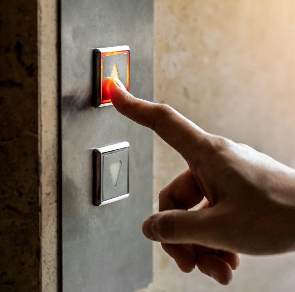 Nahaufnahme von einer Hand. An einer Wand ist das Schaltpanel eines Aufzuges zu sehen. Dieses hat zwei Knöpfe mit jeweils einem Pfeil. Der eine zeigt nach oben, der andere nach unten. Die Hand ist in Richtung dieses Panels ausgestreckt. Der Zeigefinger berührt den oberen Knopf.