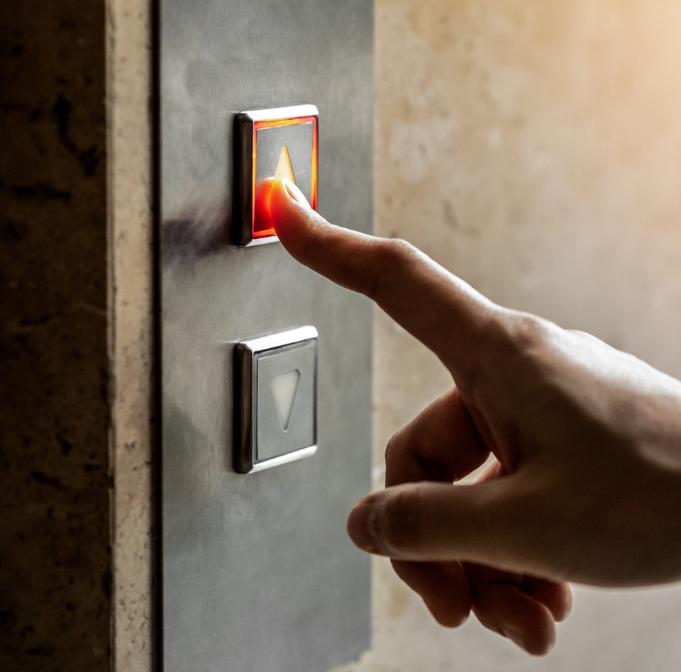 Nahaufnahme von einer Hand. An einer Wand ist das Schaltpanel eines Aufzuges zu sehen. Dieses hat zwei Knöpfe mit jeweils einem Pfeil. Der eine zeigt nach oben, der andere nach unten. Die Hand ist in Richtung dieses Panels ausgestreckt. Der Zeigefinger berührt den oberen Knopf.