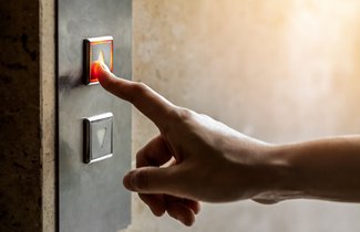 Nahaufnahme von einer Hand. An einer Wand ist das Schaltpanel eines Aufzuges zu sehen. Dieses hat zwei Knöpfe mit jeweils einem Pfeil. Der eine zeigt nach oben, der andere nach unten. Die Hand ist in Richtung dieses Panels ausgestreckt. Der Zeigefinger berührt den oberen Knopf.