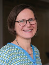 Eine Frau mit braunen Haaren lächelt freundlich in die Kamera. Sie trägt eine bunte Bluse und braune Brille. 