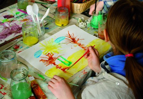 Et barn maler et billede med forskellige farver og pensler. Billedet viser palmer på stranden. På bordet står farvepotter og andet malerudstyr.