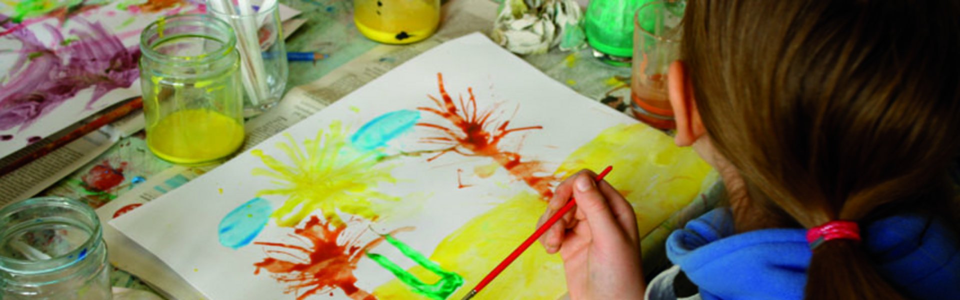 Ein Kind malt mit verschiedenen Farben und Pinseln ein Bild. Das Bild zeigt Palmen am Strand. Auf dem Tisch stehen Farbtöpfe und weitere Malutensilien.