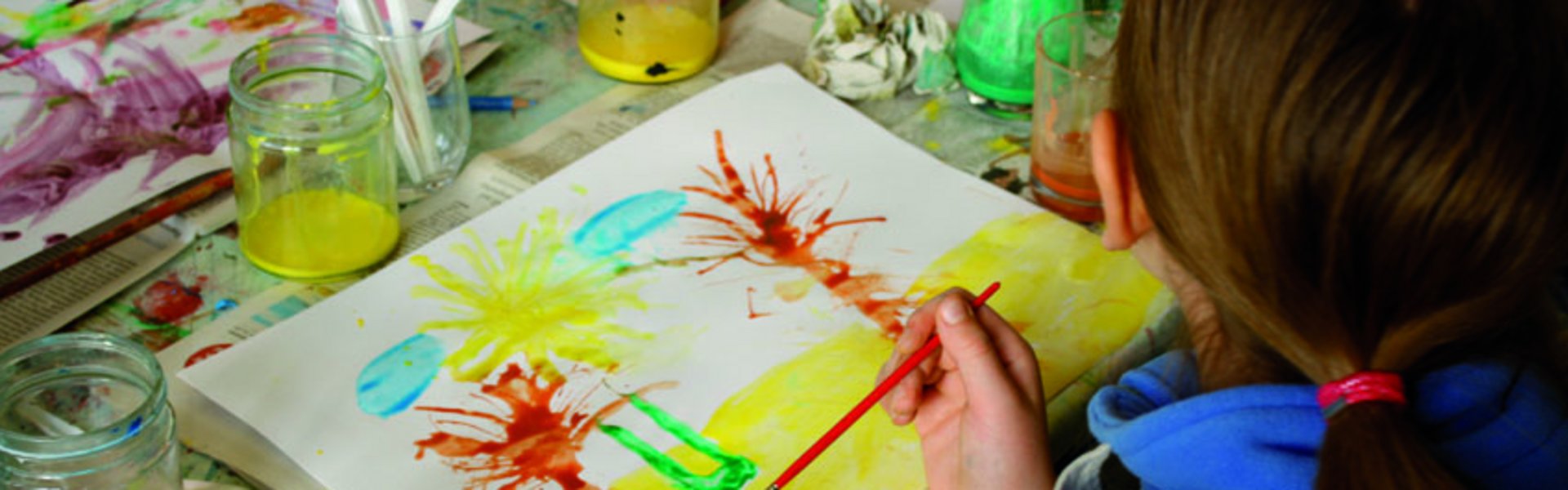 Et barn maler et billede med forskellige farver og pensler. Billedet viser palmer på stranden. På bordet står farvepotter og andet malerudstyr.
