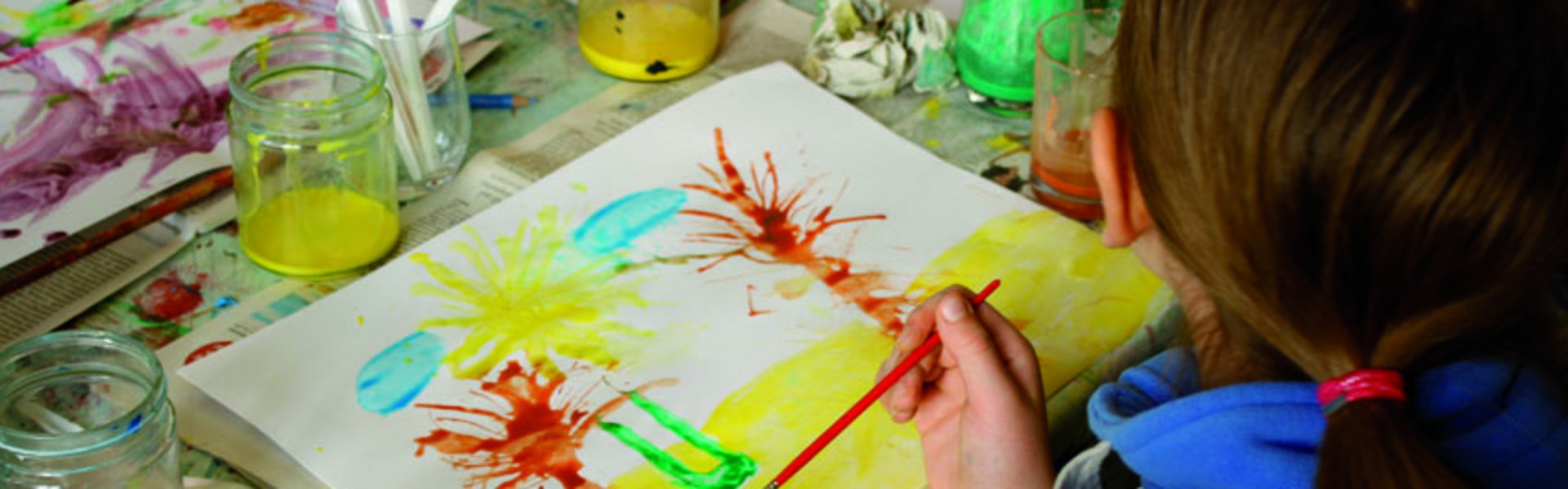 Ein Kind malt mit verschiedenen Farben und Pinseln ein Bild. Das Bild zeigt Palmen am Strand. Auf dem Tisch stehen Farbtöpfe und weitere Malutensilien.