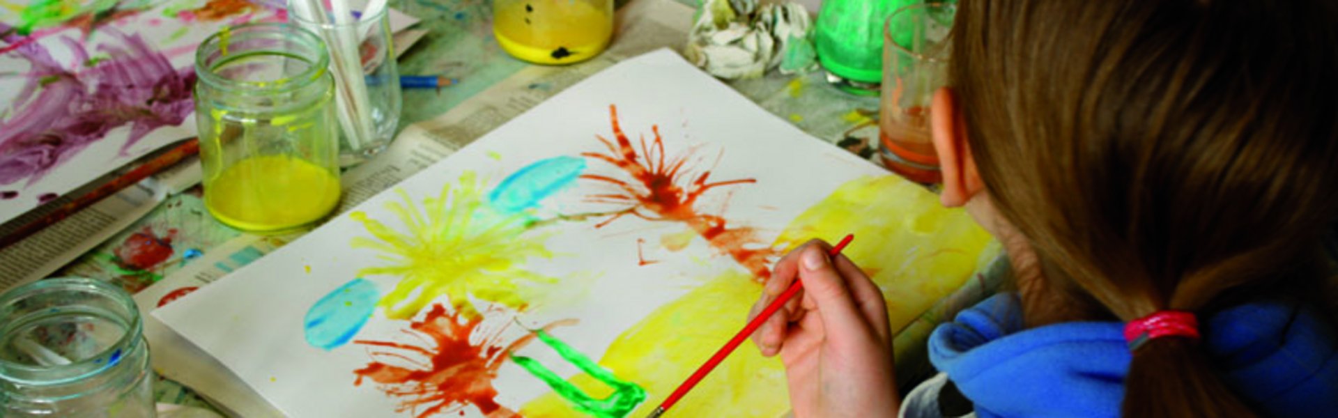 Et barn maler et billede med forskellige farver og pensler. Billedet viser palmer på stranden. På bordet står farvepotter og andet malerudstyr.