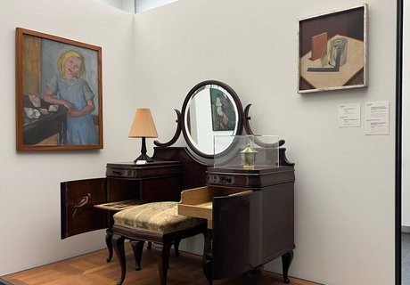 Eine bunte Fotografie. Darauf zu sehen ist im Vordergrund ein kleines Podest mit einer Rückwand und einer Seitenwand. An der Seitenwand hängt das Gemälde eines Mädchens mit blondem Haar. Sie trägt ein blaues Trägerkleid mit einem grünlich-blauem, kurzärmeligen Kragenhemd darunter. Sie lehnt mit ihrem rechten Arm auf einem Tisch. Auf dem Tisch liegen verschieden große und farbige Schneckenhäuser. Auf der Rückwand hängt in der oberen rechten Ecke ein Gemälde, auf dem verschiedene Gegenstände gezeigt werden. Auf dem Podest steht ein alter Schminktisch aus dunklem Holz. In der Mitte ist ein Spiegel. Davor steht ein kleiner Hocker aus dem gleichen Holz mit einem grün verzierten Polster. Im Hintergrund ist eine Wand zu sehen. Dort hängt das kleine Portrait eines Manns, der in seiner Hand eine Pfeife hält. Er trägt eine Schiebermütze und einen Anzug mit Krawatte. 