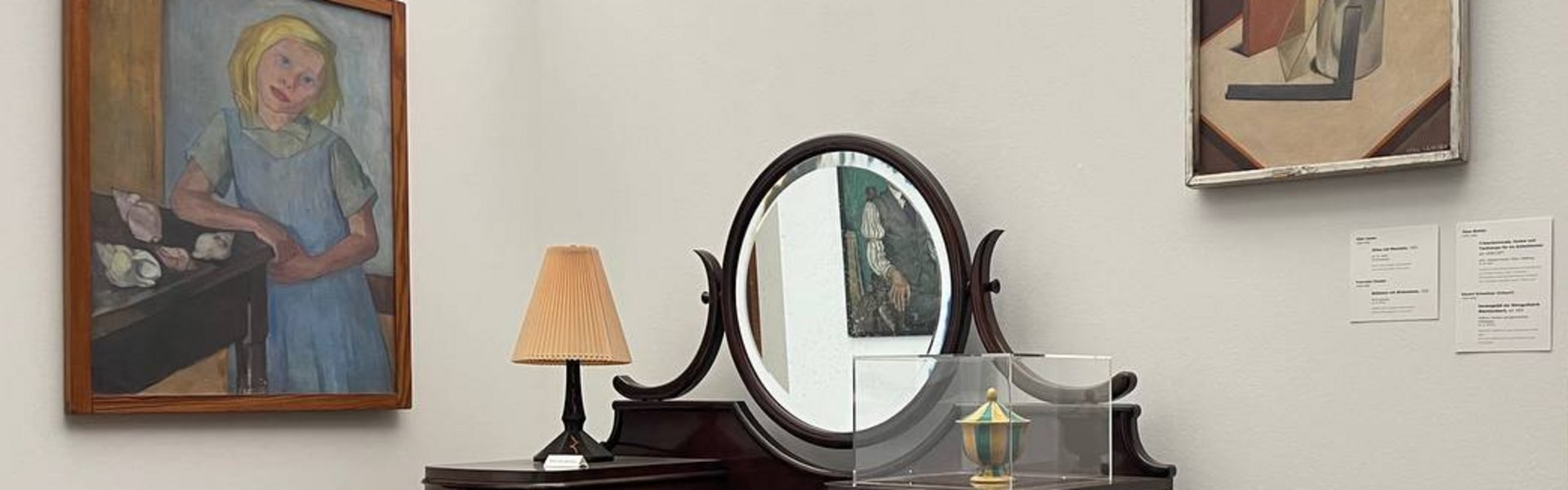 Eine bunte Fotografie. Darauf zu sehen ist im Vordergrund ein kleines Podest mit einer Rückwand und einer Seitenwand. An der Seitenwand hängt das Gemälde eines Mädchens mit blondem Haar. Sie trägt ein blaues Trägerkleid mit einem grünlich-blauem, kurzärmeligen Kragenhemd darunter. Sie lehnt mit ihrem rechten Arm auf einem Tisch. Auf dem Tisch liegen verschieden große und farbige Schneckenhäuser. Auf der Rückwand hängt in der oberen rechten Ecke ein Gemälde, auf dem verschiedene Gegenstände gezeigt werden. Auf dem Podest steht ein alter Schminktisch aus dunklem Holz. In der Mitte ist ein Spiegel. Davor steht ein kleiner Hocker aus dem gleichen Holz mit einem grün verzierten Polster. Im Hintergrund ist eine Wand zu sehen. Dort hängt das kleine Portrait eines Manns, der in seiner Hand eine Pfeife hält. Er trägt eine Schiebermütze und einen Anzug mit Krawatte. 