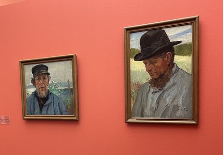 Auf dem Foto sind rechts zwei Gemälde mit Portraits eines Mannes zu sehen. Der Mann trägt eine dunkle Mütze und ein stabiles blaues Hemd. Er hat einen Bart. An der linken Wand hängen zwei weitere Bilder.