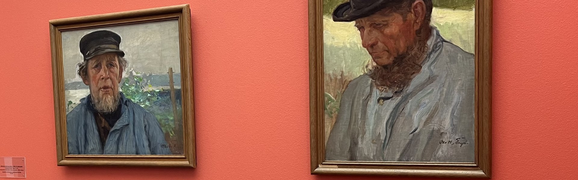 Auf dem Foto sehen Sie rechts zwei Gemälde. Beide zeigen ein Porträt von einem Mann. Ein Porträt ist ein Bild von einer Person. Der Mann trägt eine dunkle Mütze und ein stabiles blaues Hemd. Er hat einen Bart. An der linken Wand hängen zwei weitere Bilder.