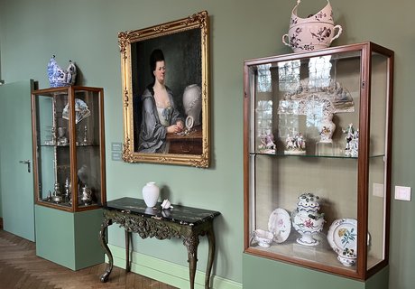 Die Fotografie zeigt in der Mitte ein Gemälde, auf dem eine Frau zu sehen ist. Das Brustbild zeigt eine Adelige im Morgenmantel. Vor ihr steht eine Vase. Unter dem Gemälde steht ein kleiner Tisch. Auf dem Tisch steht auch eine Vase. Rechts und links daneben stehen Glasvitrinen. In ihnen befinden sich verschiedene Objekte.  