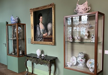 Die Fotografie zeigt in der Mitte ein Gemälde. Auf dem Gemälde sieht man eine Frau. Es ist ein Brustbild von einer Adligen, die einen Morgenmantel trägt. Ein Brustbild ist ein Bild, auf dem nur der obere Teil einer Person zu sehen ist. Vor ihr steht eine Vase. Unter dem Gemälde steht ein kleiner Tisch. Auf dem Tisch steht ebenfalls eine Vase. Rechts und links vom Tisch stehen Glasvitrinen. In diesen Vitrinen befinden sich verschiedene Objekte.