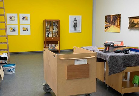 Auf dem Foto ist ein gelber Ausstellungsraum zu sehen. An der gelben Wand hängen unterschiedliche Bilder und in der Mitte steht ein Vitrinenschrank. Im Vordergrunde steht ein kleiner Transportwagen und daneben ein mobiler Arbeitstisch mit unterschiedlichen Werkzeugen. Links im Bild ist eine Holzleiter und verschiedene Malermaterialien. 