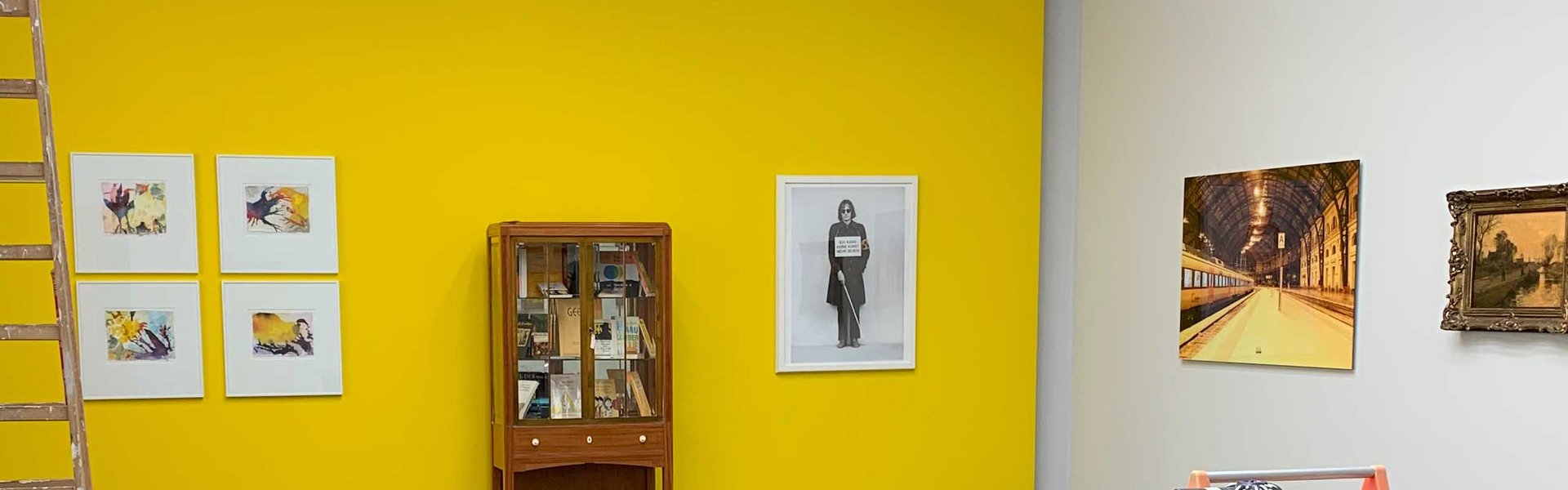 Auf dem Foto ist ein gelber Ausstellungsraum zu sehen. An der gelben Wand hängen unterschiedliche Bilder und in der Mitte steht ein Vitrinenschrank. Im Vordergrunde steht ein kleiner Transportwagen und daneben ein mobiler Arbeitstisch mit unterschiedlichen Werkzeugen. Links im Bild ist eine Holzleiter und verschiedene Malermaterialien. 