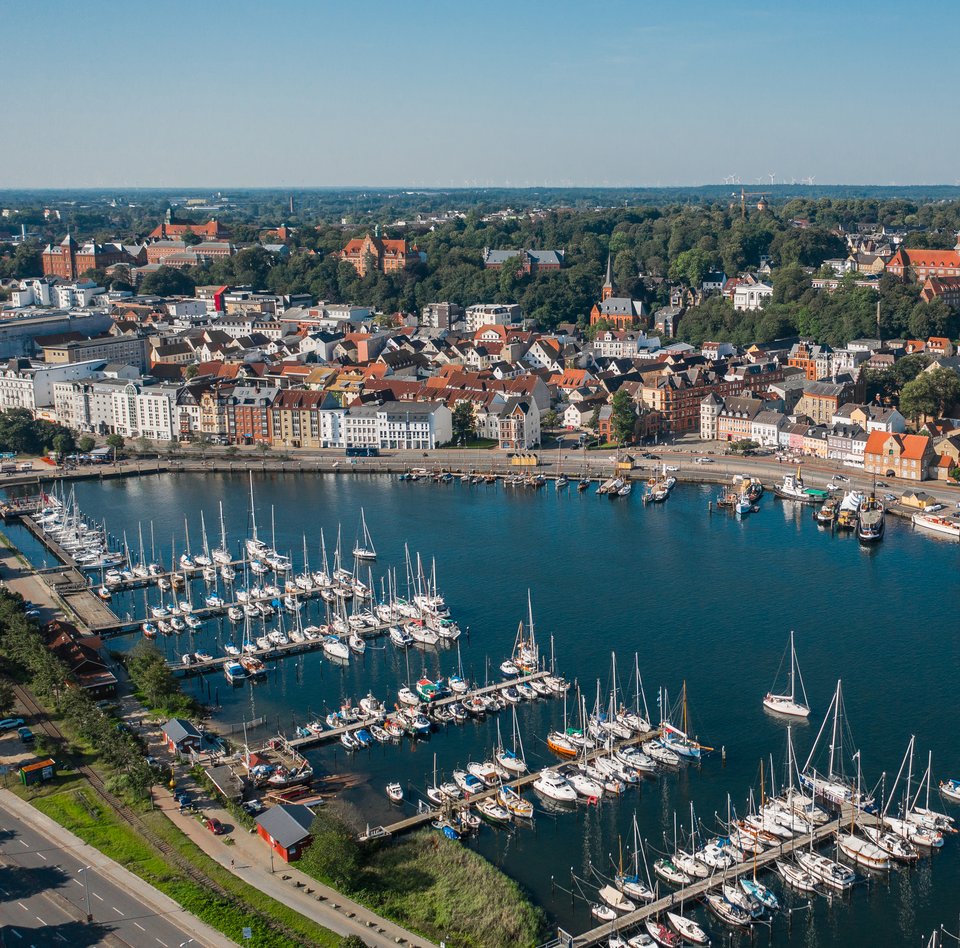 Farbige Luftaufnahme der Flensburger Innenförde. Beide Uferseiten sind bebaut und verfügen über Anlegemöglichkeiten für moderne und historische Schiffe.