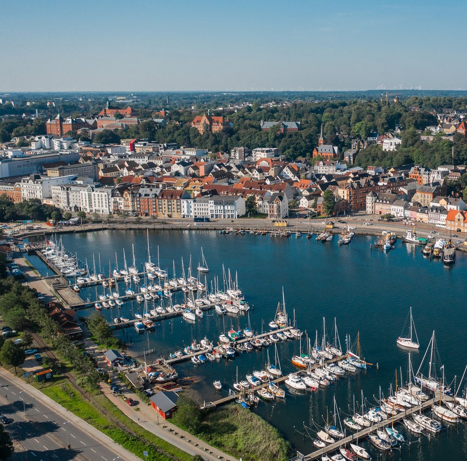 Farbige Luftaufnahme der Flensburger Innenförde. Beide Uferseiten sind bebaut und verfügen über Anlegemöglichkeiten für moderne und historische Schiffe.