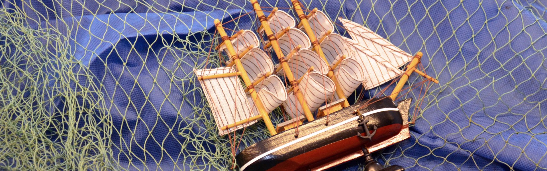 På det farvefotografi ses et blåt stykke stof med et grønt fiskenet ovenpå. Ovenpå ligger en model af et sejlskib på siden.