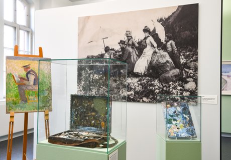 Ein großes schwarz-weiß Foto hängt an der Wand. Davor steht ein Gemälde auf einem Ständer. Daneben sind alte Pinsel und Farbe in einem Glaskasten.