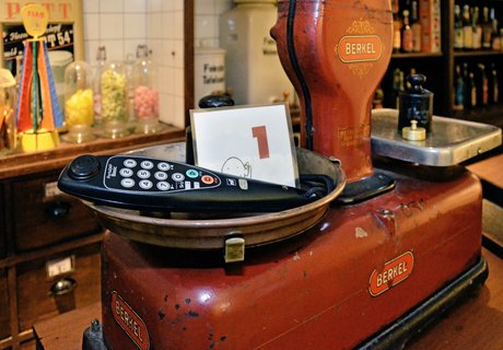 Eine rote Tafelwaage steht auf einem Tresen. Im Hintergrund sind Regale mit Süßigkeiten und Rumflaschen zu sehen. In einer der Schalen der Waage liegt ein Kopfhörer und ein kleiner Aufsteller mit der Zahl 1. 