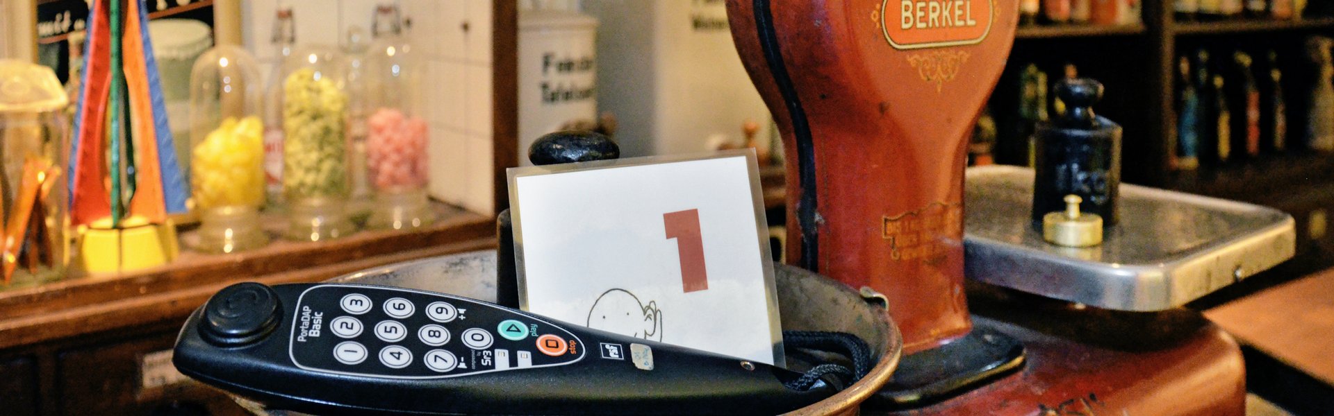 Eine rote Tafelwaage steht auf einem Tresen. Im Hintergrund sind Regale mit Süßigkeiten und Rumflaschen zu sehen. In einer der Schalen der Waage liegt ein Kopfhörer und ein kleiner Aufsteller mit der Zahl 1. 