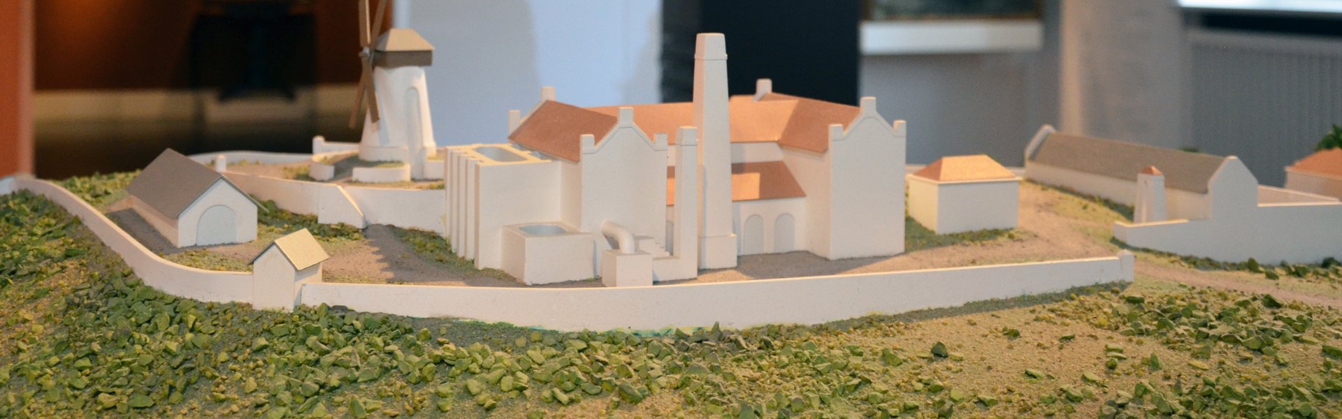 [Translate to Leichte Sprache:] Die farbige Fotografie zeigt eine Zuckerrohrplantage. Verschwommen dahinter kann man eine brauen Säule mit einer Glasabdeckung sehen. Unter dieser ist eine geschnitzte, gefesselte Figur zu sehen, dahinter ist an der Wand eine Landkarte abgebildet. Sie zeigt den Dreieckshandel. Ganz im Hintergrund ist ein weiterer Raum zu sehen mit Gemälden von Schiffen und einem Portrait zu sehen.