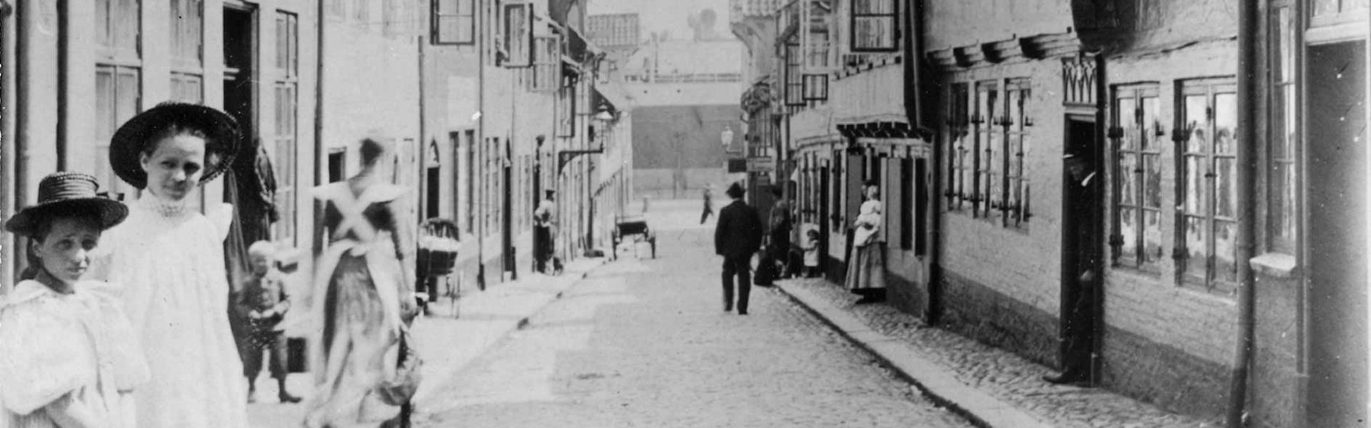 Gammelt fotografi af Oluf-Samson-gangen. I forgrunden til venstre står to piger i fine kjoler. De har begge hat på. Billedet er taget på en sådan måde, at gaden ender lige i midten af billedet. Der kan man se Flensborgs Havn.