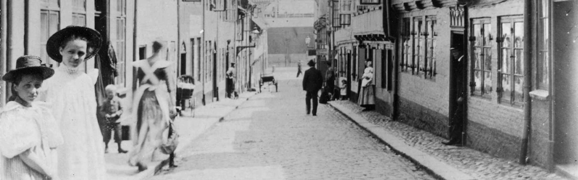 Gammelt fotografi af Oluf-Samson-gangen. I forgrunden til venstre står to piger i fine kjoler. De har begge hat på. Billedet er taget på en sådan måde, at gaden ender lige i midten af billedet. Der kan man se Flensborgs Havn.