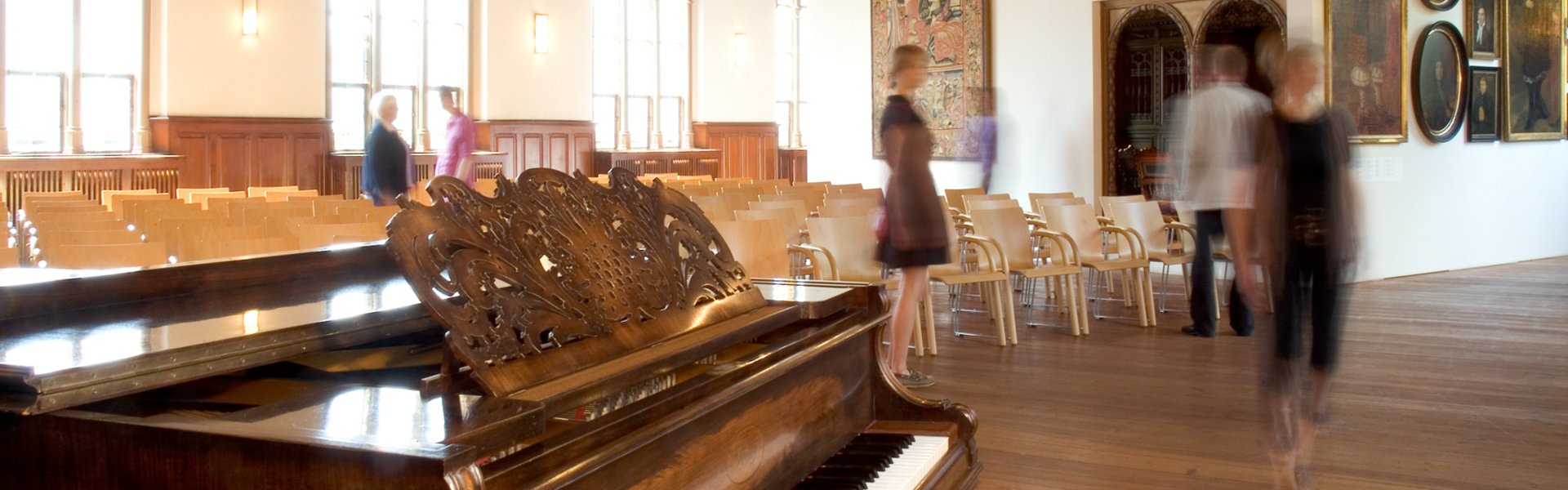 Det er auditoriet på Museumsberg Flensburg. Der kan man afholde koncerter. Der afholdes også andre arrangementer, f.eks. når der er en ny udstilling, der skal fejres. Der er mange stole og mennesker, der bevæger sig rundt. Foran ses et klaver. I baggrunden ses det parisiske værelse. Det er et rum, der er indrettet som et kunstværk.