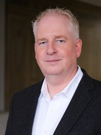 Portrait eines Mannes. Er trägt ein weißes Hemd und eine schwarze Anzugjacke.