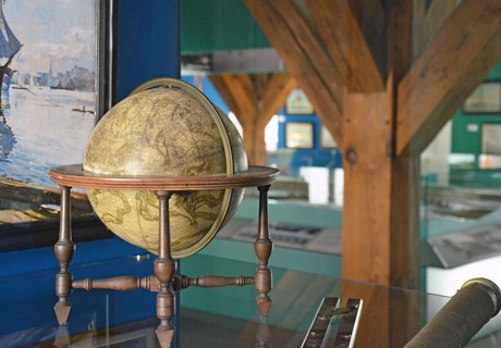 Das Farbfoto zeigt einen Globus und ein Fernrohr, das in einer Vitrine liegen. An der blauen Vitrinenwand hängt ein Gemälde, das ein Schiff zeigt. Hinter der Vitrine kann man das Stadtmodell und weitere Bilder, die an Wänden hängen sehen.