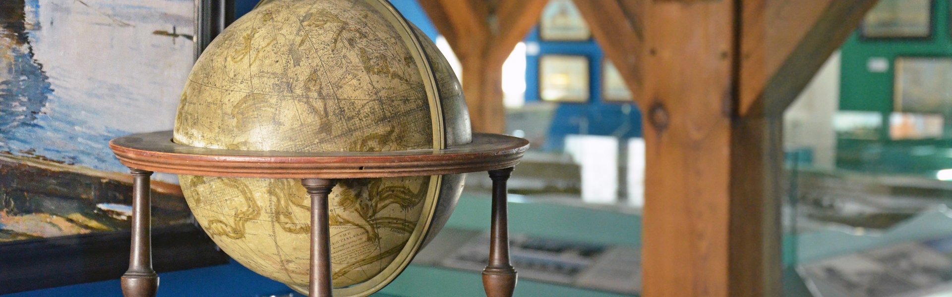 Das Farbfoto zeigt einen Globus und ein Fernrohr, das in einer Vitrine liegen. An der blauen Vitrinenwand hängt ein Gemälde, das ein Schiff zeigt. Hinter der Vitrine kann man das Stadtmodell und weitere Bilder, die an Wänden hängen sehen.