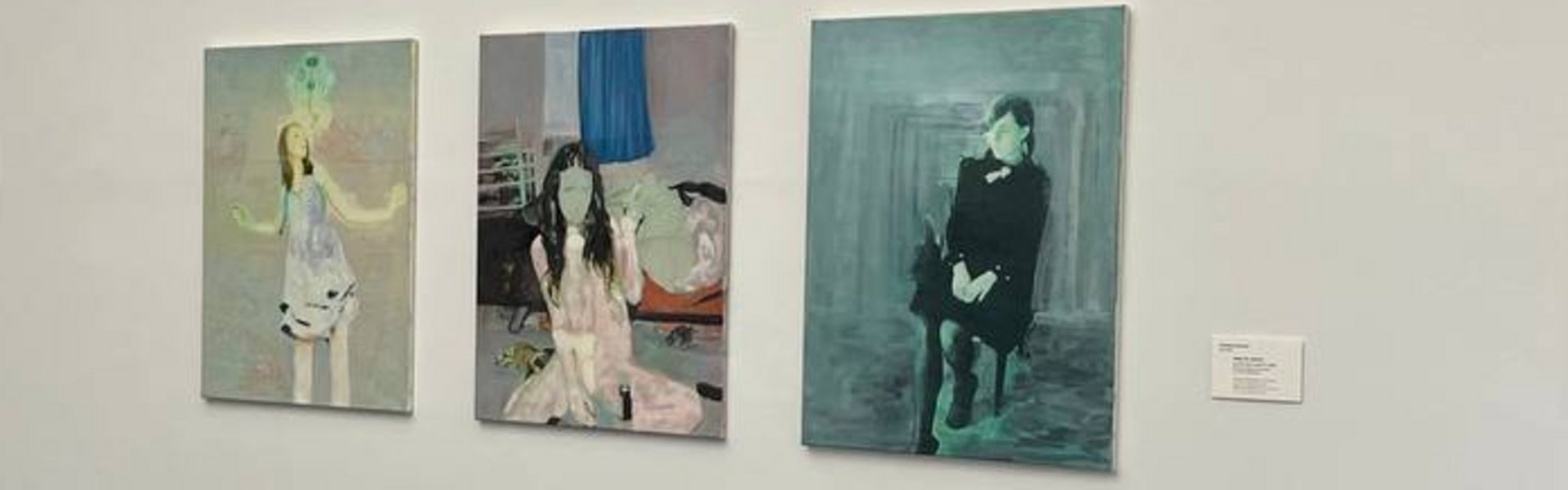 Auf der bunten Fotografie sieht man einen Ausstellungsraum mit weißen Wänden. An einer Wand hängen verschiedene Portraits von jungen Frauen. Hinten sieht man einen Flur. An der weißen Wand dort hängt eine Fotografie.