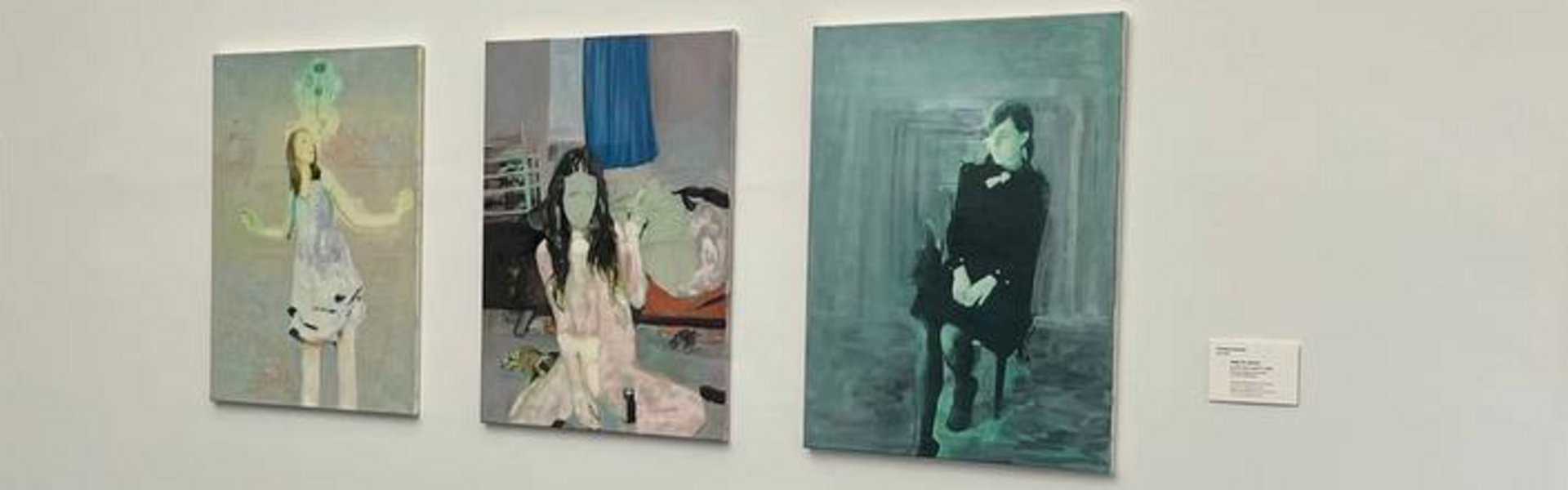 Eine bunte Fotografie. Darauf ist ein Ausstellungsraum mit weißen Wänden zu sehen. An einer der Wände hängen verschiedene Potraits von jungen Frauen. Im Hintergrund ist der Teil eines Flures zu sehen. Dort hängt an der weißen Wand eine Fotografie.