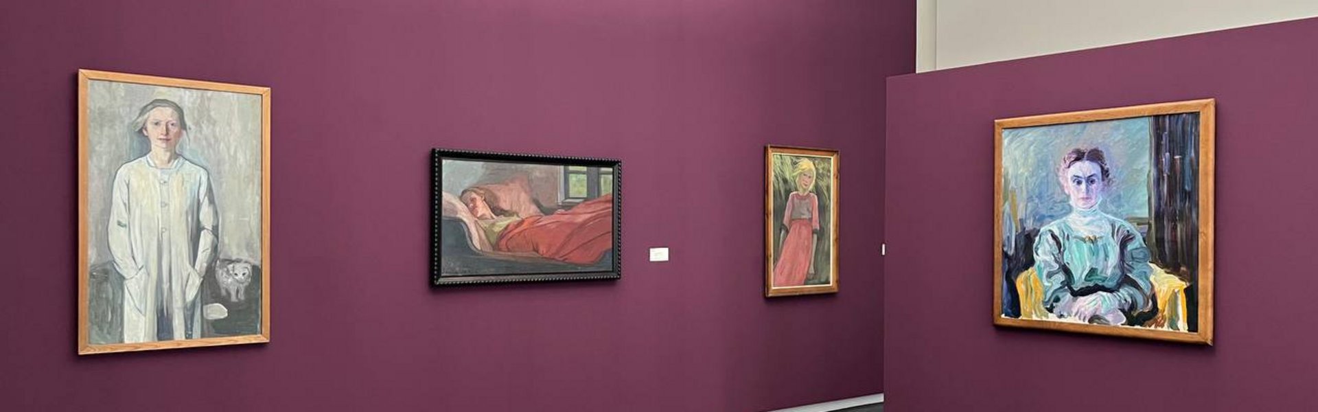 Eine bunte Fotografie. Darauf sind Teile eines Ausstellungsraums des Museumsberg Flensburgs zu sehen. Die Wände sind in einem Fuchsia-Ton gestrichen. An der einen Wand hängen drei Gemälde der Künstlerin Käte Lassen. Auf den drei Gemälden sind unterschiedliche Frauen dargestellt. Auf der Wand rechts befindet sich ein weiteres Portrait von einem anderen Künstler. 