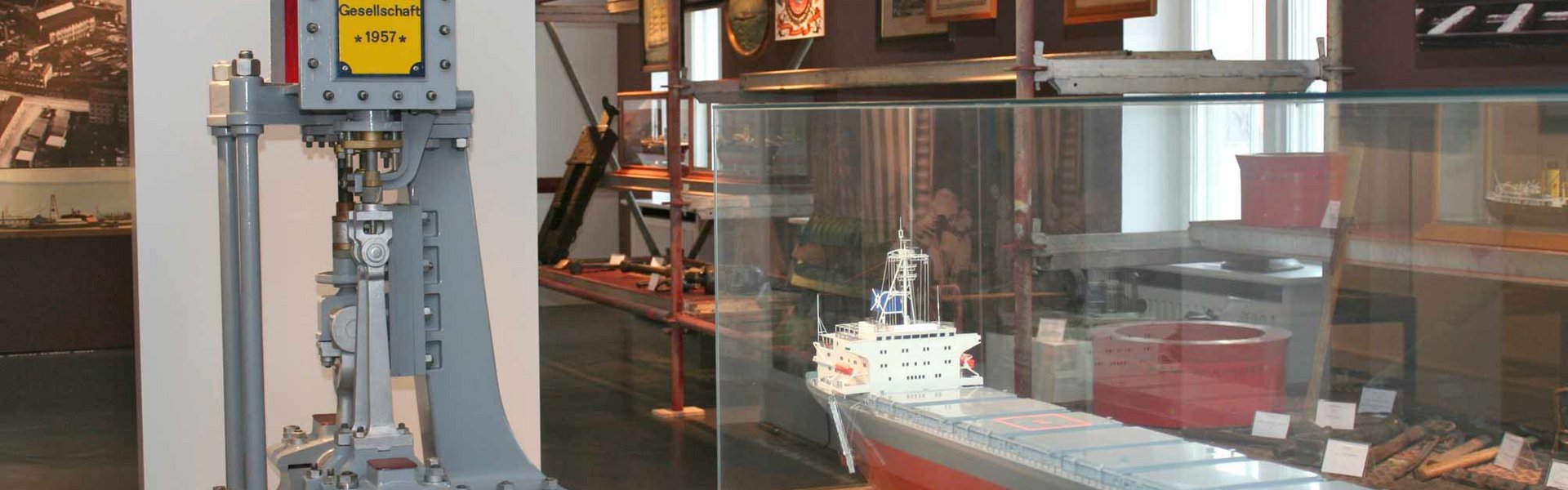 Mittig im Raum steht ein Schiffsmodell eines Stahlschiffes in einer Vitrine. Dahinter eine Maschine der Flensburger Schiffsbau Gesellschaft. Über die komplette rechte Seite des Raumes steht ein Stahlregal mit unterschiedlichen Werkzeugen die u.a. zum Bau eines Schiffes erforderlich sind.