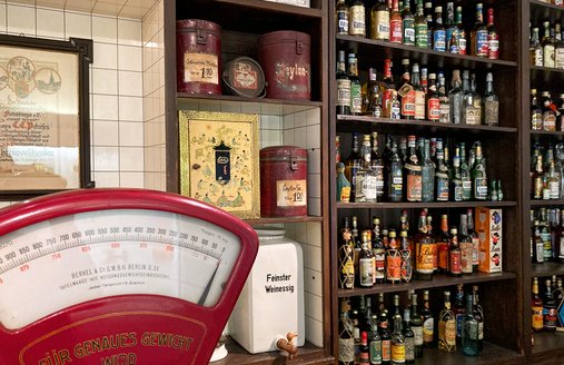 På billedet ses en kolonialvarebutik. Helt til venstre på disken står en rød vægt, bagved er der en reol med utallige romflasker samt te- og kaffedåser.