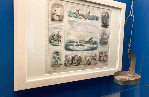 I en vitrine med blå bagvæg hænger en tegning med flere billeder om emnet hvalfangst. Til højre for den hænger en gammel lampe, der blev fyret med tran.