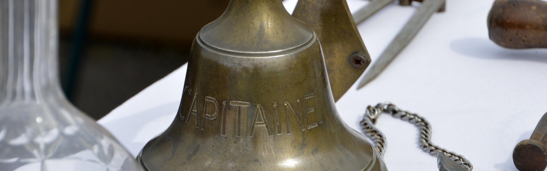 Auf diesem Bild ist eine Schiffsglocke. Sie ist aus Messing. Sie steht auf einem Tisch. Neben der Glocke liegen andere maritime Gegenstände.