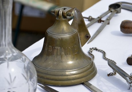 Auf diesem Bild ist eine Schiffsglocke. Sie ist aus Messing. Sie steht auf einem Tisch. Neben der Glocke liegen andere maritime Gegenstände.