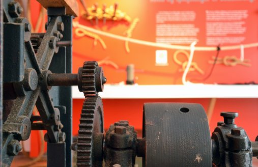 Im Vordergrund steht eine alte Maschine. Sie heißt Reep-Schlag-Maschine. Sie ist aus mehreren Zahn-Rädern und einer Kurbel hergestellt. Davor ist ein Seil gespannt, an das kleine Seile befestigt sind. So macht man Seile.