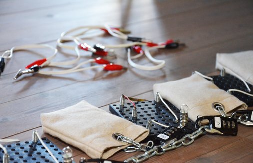 Auf dem Foto ist eine Gliederkette aus Metall zu sehen. Sie liegt auf einem Dielenboden. An der Kette sind kleine Säckchen mit jeweils einem Zahlenschloss gekettet. Zwischen jedem Säckchen ist eine Morsestation mit einem Zahlenschloss an die Kette gekettet. Im Hintergrund leicht verschwommen kann man Kabel mit Krokodilsteckern erkennen.