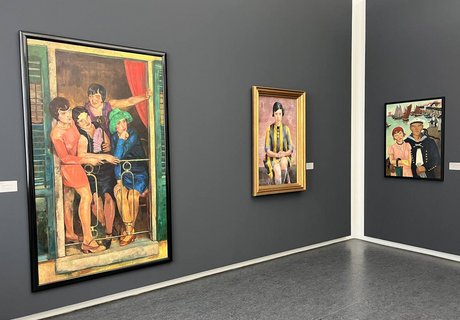 Das Foto zeigt eine Ecke eines Ausstellungsraums. Die Wände sind in einem dunklen Grau gestrichen. An der linken Wand hängen zwei Gemälde. Und an der rechten Wand hängt ein weiteres Gemälde. Das vorderste Gemälde zeigt vier Frauen. Sie tragen Kleider im Stil der 1920er Jahre. Die Frauen stehen auf einem kleinen Balkon und schauen etwas rechts unter sich an. Rechts neben diesem Gemälde ist ein weiteres zu sehen. Es zeigt eine junge Frau, die sitzt und den Betrachter aus dem Bild anschaut. Sie trägt ein gelb-schwarz gestreiftes Kleid. Das nächste Gemälde zeigt eine Frau, die leicht von einem Mann umarmt wird. Die Frau trägt ein rosa Kleid, eine rote Mütze, eine kleine braune Handtasche und einen grünen Regenschirm. Der hängt an ihrem rechten Arm. Der Mann trägt eine Matrosenuniform. Er hat den rechten Arm um die Frau gelegt und hält in der linken Hand weiße Handschuhe. Hinten in diesem Bild sieht man einen Teil eines Hafens mit Holzstegen. Auf dem Wasser sind einige Segelboote und andere Boote zu sehen.