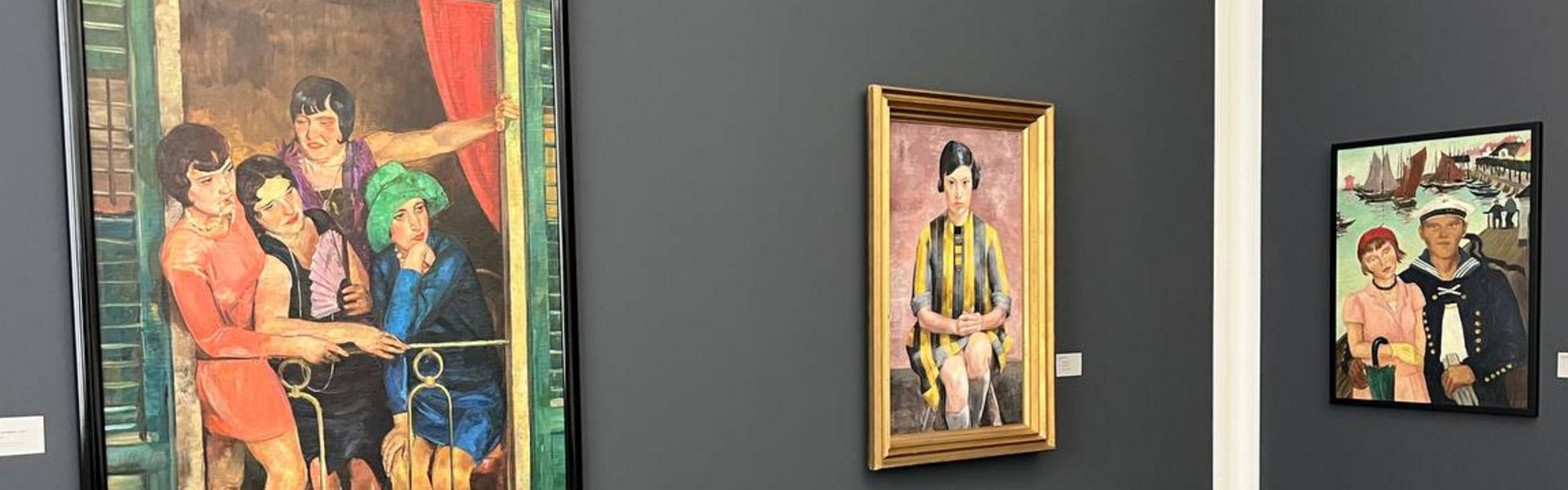 Eine bunte Fotografie. Man sieht eine Ecke eines Ausstellungsraums. Die Wände sind in einem dunklen grau gestrichen. An der linken Wand sieht man zwei Gemälde hängen und an der rechten Wand ist ein weiteres Gemälde zu sehen. Das Vorderste ist ein buntes Gemälde, auf dem vier Frauen in Kleidern im Stile der 1920er Jahre gekleidet sind.  Sie stehen auf einem kleinen Balkon und schauen sich etwas rechts unter ihnen an. Rechts daneben ist das Gemälde einer Frau zu sehen. Sie ist sitzend dargestellt und schaut den Betrachter aus dem Bild an. Sie trägt ein gelb-schwarz gestreiftes Kleid. Das Gemälde an der Wand daneben zeigt eine Frau, die leicht von einem Mann umarmt wird. Die Frau trägt ein rosa Kleid, eine rote Mütze, eine kleine braune Handtasche und einen grünen Regenschirm, der geschlossen an ihrem rechten Arm hängt. Der Mann trägt eine Matrosenuniform. Er hat den rechten Arm um die Frau gelegt und in der linken Hand hält er weiße Handschuhe. Im Hintergrund sieht man den Abschnitt eines Hafens mit seinen Holzstegen. Auf dem Wasser in der linken Hälfte des Hintergrunds, sind ein paar Segelboote und andere Boote zu sehen.