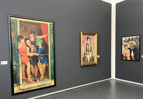 Eine bunte Fotografie. Man sieht eine Ecke eines Ausstellungsraums. Die Wände sind in einem dunklen grau gestrichen. An der linken Wand sieht man zwei Gemälde hängen und an der rechten Wand ist ein weiteres Gemälde zu sehen. Das Vorderste ist ein buntes Gemälde, auf dem vier Frauen in Kleidern im Stile der 1920er Jahre gekleidet sind.  Sie stehen auf einem kleinen Balkon und schauen sich etwas rechts unter ihnen an. Rechts daneben ist das Gemälde einer Frau zu sehen. Sie ist sitzend dargestellt und schaut den Betrachter aus dem Bild an. Sie trägt ein gelb-schwarz gestreiftes Kleid. Das Gemälde an der Wand daneben zeigt eine Frau, die leicht von einem Mann umarmt wird. Die Frau trägt ein rosa Kleid, eine rote Mütze, eine kleine braune Handtasche und einen grünen Regenschirm, der geschlossen an ihrem rechten Arm hängt. Der Mann trägt eine Matrosenuniform. Er hat den rechten Arm um die Frau gelegt und in der linken Hand hält er weiße Handschuhe. Im Hintergrund sieht man den Abschnitt eines Hafens mit seinen Holzstegen. Auf dem Wasser in der linken Hälfte des Hintergrunds, sind ein paar Segelboote und andere Boote zu sehen.