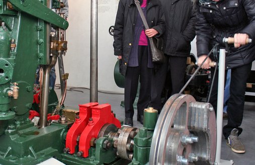 I forgrunden ses en maskine, som er udstyret med en drevmekanisme, der kan betjenes manuelt af museets gæster. En pige sætter maskinen i gang. To andre personer ser på.