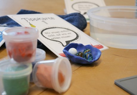 Eine bunte Fotografie. Darauf zu sehen sind verschiedene Gegenstände. Ganz links im Vordergrund befinden sich vier kleine Plastikdöschen. Diese sind durchsichtig. Jedes Döschen hat unterschiedlich bunte Knetmasse enthalten. In der Mitte des Fotos liegt eine Papierkarte, auf der „Tippkarte 1“ steht. Auf dem untern Teil dieser Karte liegt eine Schale geformt aus blauer Knete. Darin befinden sich vier Murmeln. Im Hintergrund steht eine durchsichte Plastikschüssel mit klarem Wasser darin. Daneben und halb unter der Karte liegt ein blaues Handtuch. 