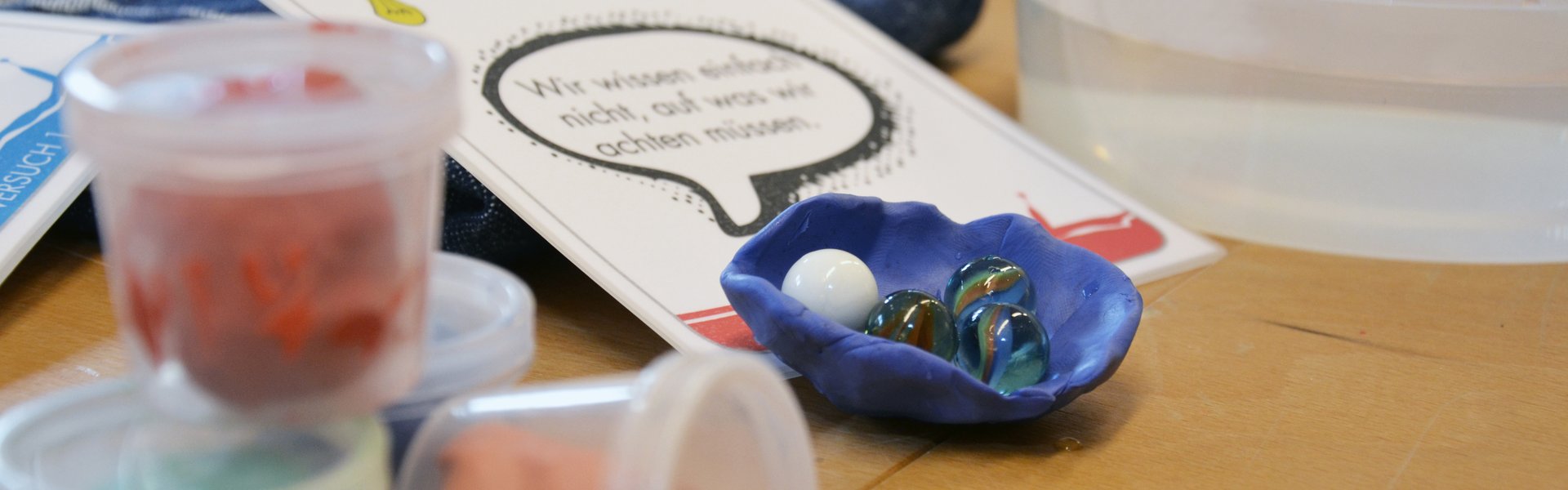 Eine bunte Fotografie. Darauf zu sehen sind verschiedene Gegenstände. Ganz links im Vordergrund befinden sich vier kleine Plastikdöschen. Diese sind durchsichtig. Jedes Döschen hat unterschiedlich bunte Knetmasse enthalten. In der Mitte des Fotos liegt eine Papierkarte, auf der „Tippkarte 1“ steht. Auf dem untern Teil dieser Karte liegt eine Schale geformt aus blauer Knete. Darin befinden sich vier Murmeln. Im Hintergrund steht eine durchsichte Plastikschüssel mit klarem Wasser darin. Daneben und halb unter der Karte liegt ein blaues Handtuch. 