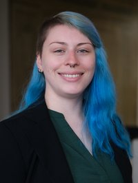 Portrait einer jungen Frau. Sie trägt dunkle Kleidung und hat blau gefärbte Haare.