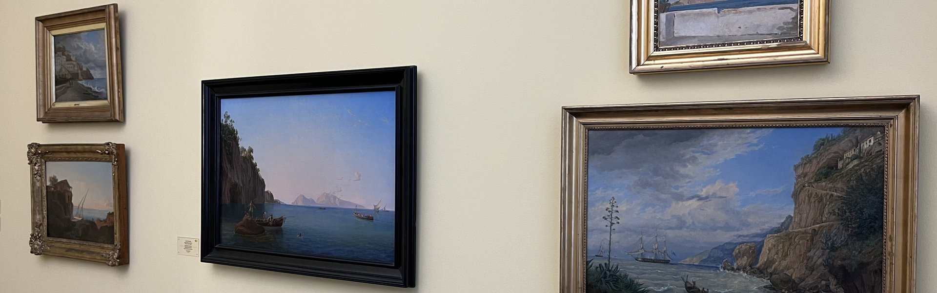 Das Foto zeigt fünf Bilder. Sie hängen an einer hellbeigen Wand. In der Mitte hängt ein Bild in einem schwarzen Rahmen. Links davon hängen zwei Bilder übereinander. Rechts davon hängen auch zwei Bilder übereinander. Auf allen Bildern sind Landschaften mit dem Meer zu sehen.