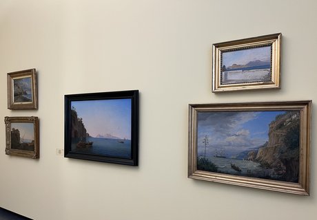 Das Foto zeigt fünf Gemälde. Die Bilder hängen vor einer hellbeigen Wand. In der Mitte hängt ein Bild mit einem schwarzen Rahmen. Links daneben hängen zwei Bilder übereinander. Rechts daneben hängen zwei Bilder übereinander. Auf allen Bildern sind Landschaften mit dem Meer abgebildet.