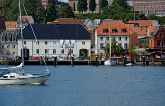 Auf dem Foto ist das Hauptgebäude des Flensburger Schifffahrtsmuseums abgebildet. Das Foto ist vom Ostufer aus aufgenommen, sodass vor dem Museum die Förde zu sehen ist. Von links kommt ein modernes Segelschiff ins Bild reingesegelt. 